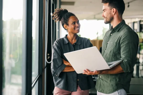 Two colleagues standing by a window, smiling and discussing work while holding an open laptop.
