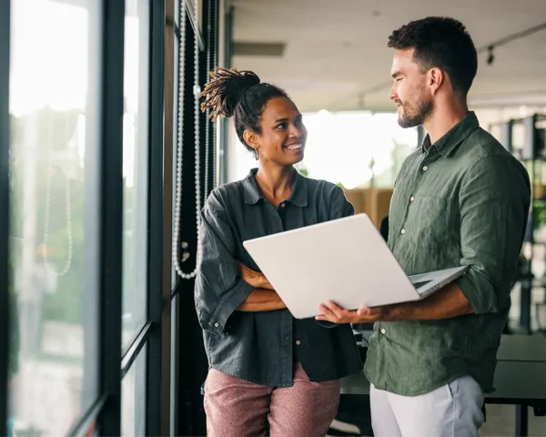 Two colleagues standing by a window, smiling and discussing work while holding an open laptop.